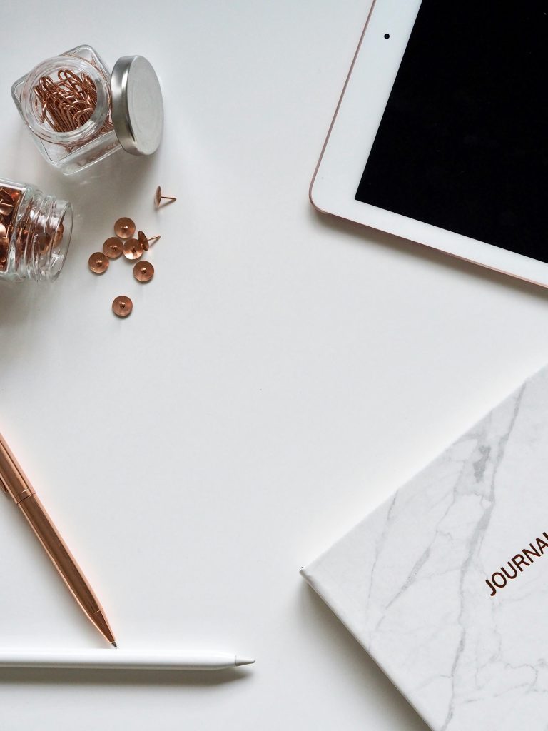 A stylish workspace featuring rose gold office supplies, a journal, and a tablet in a flat lay arrangement.